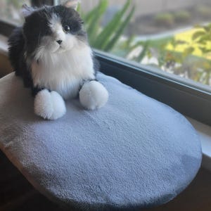 May include: A plush toy cat with black and white fur sitting on a soft, oval-shaped light gray cushion. The cat has long, fluffy fur, white paws, and dark eyes. The cushion has a smooth, velvety texture and is positioned on a flat surface.