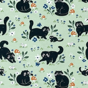 May include: A light green fabric featuring a repeating pattern of black cats in various poses, interspersed with orange mushrooms, small white and blue flowers, green leaves, blue butterflies, and tiny white birds scattered across the surface.