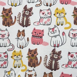 May include: A white fabric featuring a repeating pattern of cartoon cats in various colors including brown, pink, yellow, and white. The cats are depicted in different poses, some wearing collars or small neckerchiefs, with simple line-drawn facial features.