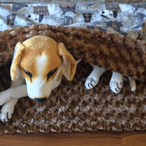 May include: A plush beagle toy resting inside a brown, textured, dimpled fabric sleeping bag. The sleeping bag features a secondary layer of light gray fabric with a repeating pattern of stylized dogs and brown plaid squares on the exterior.