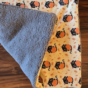 May include: A rectangular blanket featuring a cream-colored side with a repeating pattern of black cats hugging orange pumpkins and small brown leaves, and a reverse side made of thick, textured gray fleece fabric with a soft, plush appearance.