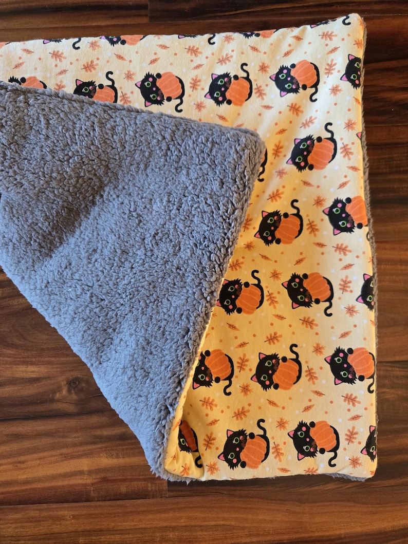 May include: A rectangular blanket featuring a cream-colored side with a repeating pattern of black cats hugging orange pumpkins and small brown leaves, and a reverse side made of thick, textured gray fleece fabric with a soft, plush appearance.