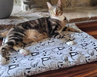 Puede incluir: Un gato atigrado marrón y negro con patas y pecho blancos tumbado sobre una alfombrilla rectangular. La alfombrilla presenta un patrón repetitivo de gatos dibujados en blanco y negro sobre un fondo gris claro con corazones.