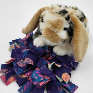 May include: A plush toy bunny with brown, black, and white fur sits atop a purple fabric mat with a colorful tie-dye pattern. The bunny has long, floppy ears and a soft, cuddly appearance. The mat is made of strips of fabric.