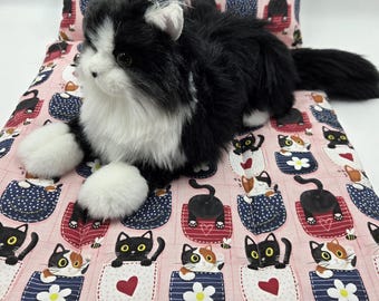 Puede incluir: Un peluche de gato de pelo largo en blanco y negro sobre un conjunto de alfombrilla y almohada rectangular. La tela presenta un patrón repetitivo de varios gatos asomándose por bolsillos coloridos con motivos de corazones y flores en rosa.