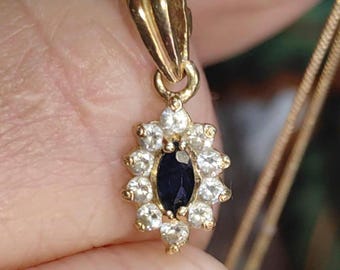 Pode incluir: Um colar de ouro com um pingente que tem uma pedra oval azul escura cercada por um halo de pedras menores e transparentes.