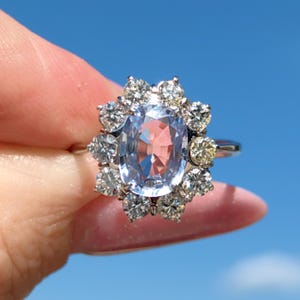 May include: A silver ring featuring a large, oval-cut, light blue gemstone surrounded by a halo of smaller, round, clear and yellow gemstones. The ring is set against a bright blue sky.