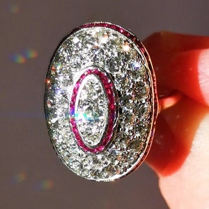 Puede incluir: Un anillo de plata con una gran piedra central de forma ovalada rodeada de diamantes más pequeños. La piedra central está rodeada por una banda de rubí rojo.