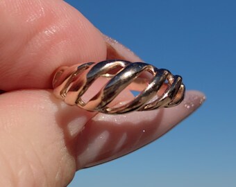 May include: A rose gold ring with a unique open, spiral design. The ring's metal has a polished finish, reflecting light. The ring is held against a clear blue sky background, highlighting its intricate form. This piece is a statement accessory.