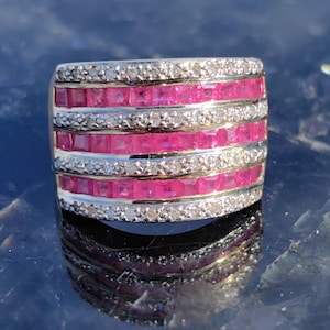May include: A wide band ring with five rows of pink square-shaped gemstones set in a gold band. The gemstones are surrounded by smaller, clear round gemstones.
