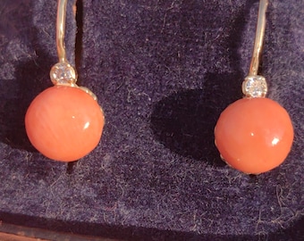 May include: A pair of gold drop earrings featuring round, coral-colored beads. Each earring has a small, clear gemstone accent above the coral bead. The earrings are displayed against a dark purple, textured background.