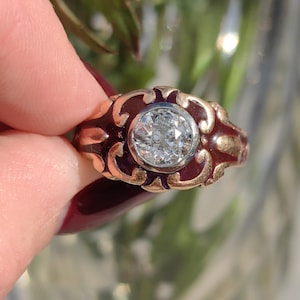 May include: A vintage gold ring with a large, sparkling diamond center stone. The ring features a dark red enamel band with ornate gold filigree detailing. The ring is held by a hand with a burgundy nail polish.