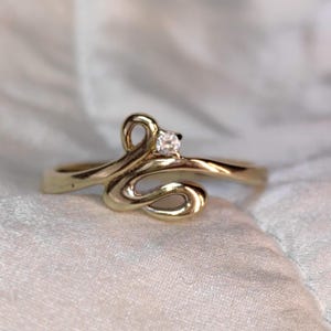 May include: A gold-toned ring with a swirling design and a small, clear gemstone. The ring's band curves into an abstract shape, with a single, round diamond set at the top. The ring is set against a soft, white background.