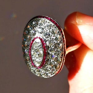 May include: An oval-shaped ring featuring a central marquise-cut diamond surrounded by a halo of small round diamonds. A thin border of vibrant red rubies encircles the center, while an outer layer of pavé-set diamonds completes the design.