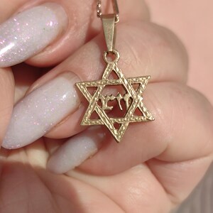 May include: Gold-colored Star of David pendant necklace. The pendant features a six-pointed star design with an inscription in the center. The necklace is displayed against a soft background.