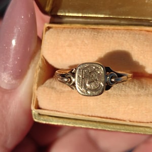 Puede incluir: Un anillo de oro con una cara cuadrada con un diseño grabado, posiblemente iniciales. El anillo se presenta en una pequeña caja abierta de color dorado con un interior color melocotón. La banda del anillo es fina y delicada.