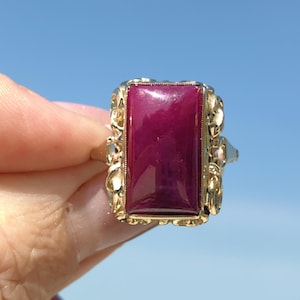 May include: A gold ring featuring a large, rectangular, deep red gemstone. The ring has an ornate, filigree-style setting with intricate details. The background is a clear, blue sky.