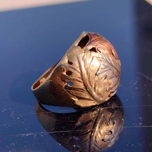 May include: A gold ring with a leaf design. The ring is made of a thick band and has a domed top. The leaf design is intricate and detailed.