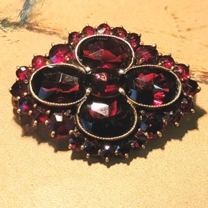 May include: A vintage brooch with a cluster of red garnet stones set in a gold tone metal. The brooch features a central design of four oval-shaped stones surrounded by smaller stones.