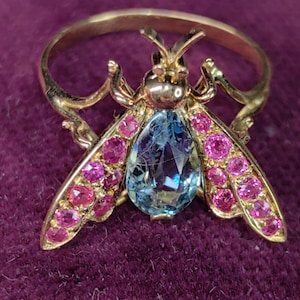 May include: A gold ring with a bee motif. The wings are set with pink gemstones, and the body features a large, light blue gemstone. The ring is displayed against a purple background.