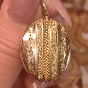 May include: A gold oval locket with detailed vertical and floral patterns. A twisted rope design runs down the centre. The locket is suspended from a small gold loop.