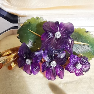 May include: A gold brooch with carved amethyst flowers and green leaves. The flowers have diamond accents.