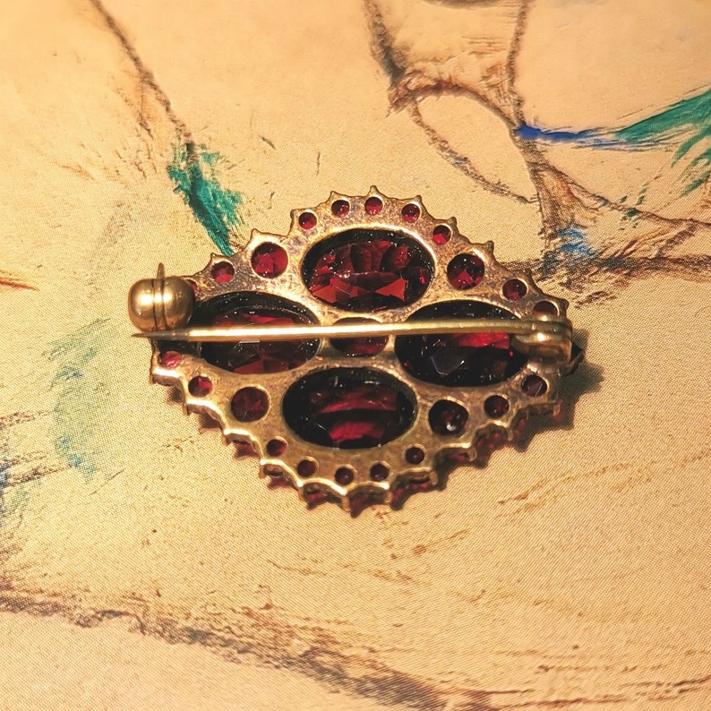 May include: A gold brooch with a red gemstone design. The brooch has a pin and catch on the back.
