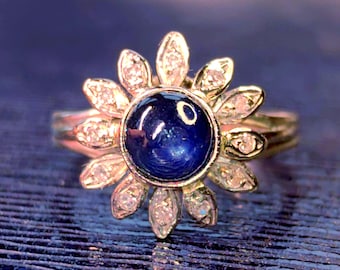 May include: A gold ring featuring a central round cabochon-cut deep blue sapphire surrounded by a halo of twelve marquise-cut clear white diamonds arranged in a flower-like pattern. The metal band is polished gold, supporting the floral setting.