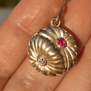 Può includere: Un ciondolo color oro con un design floreale. Il ciondolo presenta una piccola gemma rotonda e trasparente e una gemma rosa rotonda più grande. Il ciondolo è rotondo con un anello nella parte superiore per una catena.