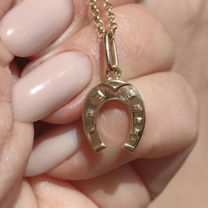 May include: A gold-colored horseshoe pendant necklace. The horseshoe charm is detailed with square and diamond-shaped accents. The necklace chain is also gold-colored and delicate. The pendant is suspended from a small loop.