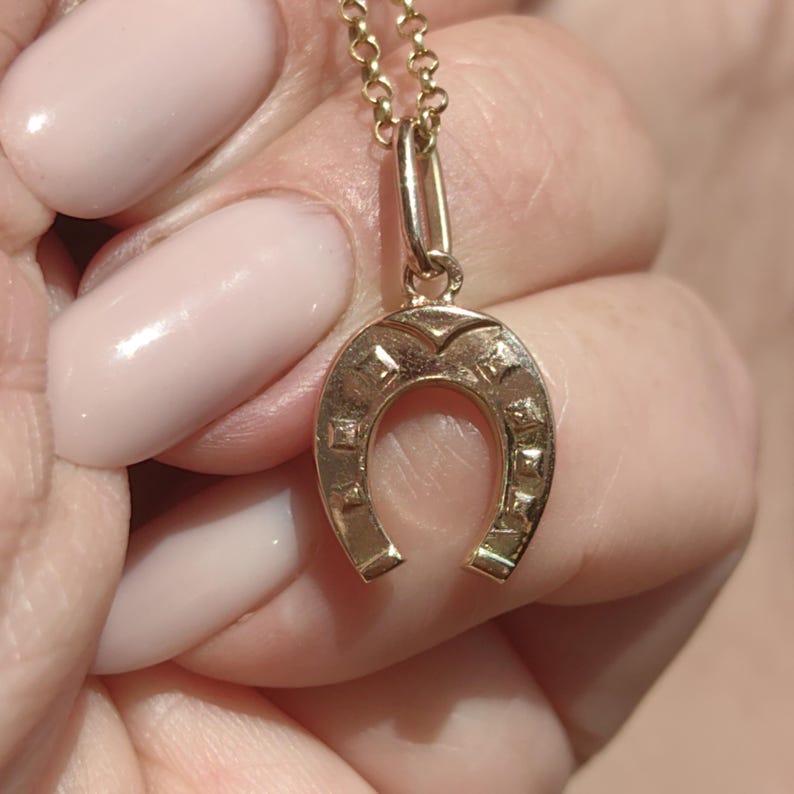 May include: A gold-colored horseshoe pendant necklace. The horseshoe charm is detailed with square and diamond-shaped accents. The necklace chain is also gold-colored and delicate. The pendant is suspended from a small loop.
