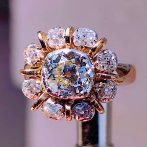 May include: A gold ring featuring a large, central diamond surrounded by smaller diamonds. The diamonds are clear and sparkling, set in a floral-like arrangement. The ring has a classic, elegant design.