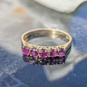 Op de afbeelding: Een gouden ring met vijf levendige roze edelstenen. De edelstenen zijn gezet in individuele prongen en de ringband is van eenvoudig, gepolijst goud. De ring wordt getoond op een reflecterend, donker oppervlak.