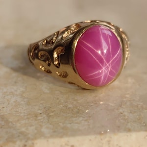 Puede incluir: Un anillo de oro con una piedra preciosa de rubí estrellado rosa. El anillo tiene un diseño detallado y adornado.