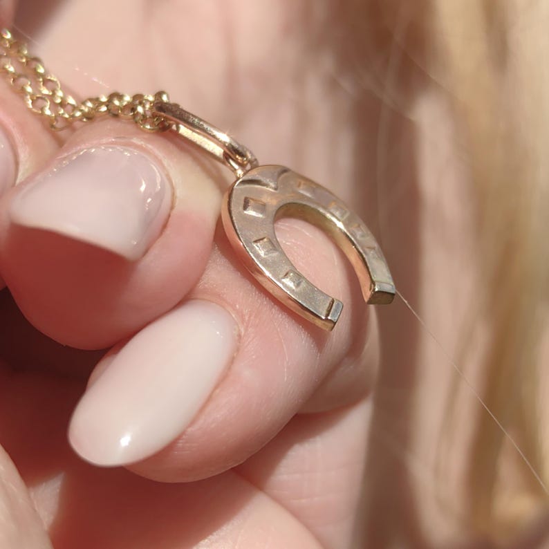 May include: A gold-colored horseshoe pendant necklace. The horseshoe charm is detailed with small square cutouts and is suspended from a delicate chain. The necklace is held in a hand with light pink nail polish.
