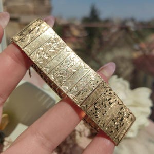Op de afbeelding: Een gouden armband met een gedetailleerd, ingewikkeld ontwerp. De armband is gemaakt van gekoppelde, rechthoekige stukken met een bloemenpatroon.
