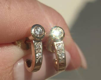 Puede incluir: Un par de pendientes de aro dorados. Cada pendiente presenta un diamante engastado en bisel en la parte superior y diamantes más pequeños a lo largo del cuerpo curvo. Los pendientes se muestran sobre un fondo borroso.