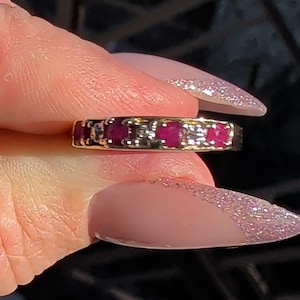 Peut inclure: Une bague en or ornée de plusieurs pierres précieuses rouges vives, taillées en carré. La conception de la bague présente des pierres rouges alternées et de plus petites pierres d'accent claires, créant un effet scintillant. La bague est tenue sur un fond flou.