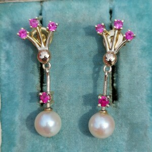 May include: A pair of gold earrings with pink gemstones and white pearls. The earrings feature a cluster of five pink gemstones at the top and a single pink gemstone below a white pearl.