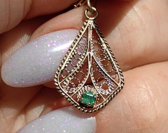 May include: A gold filigree teardrop earring with a small, square emerald gemstone. The earring features intricate openwork and a delicate design. The earring is held up by a gold hook.