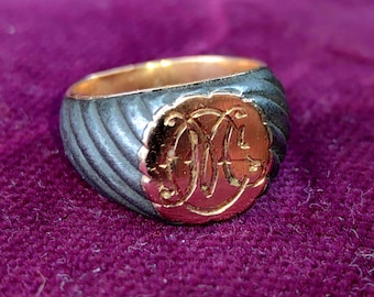 Puede incluir: Un anillo negro y dorado con una banda texturizada y un monograma circular. El monograma está grabado en oro y presenta las letras "M" y "C".