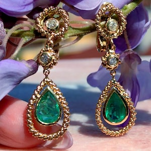 May include: Gold drop earrings with emerald-green teardrop stones. The earrings have a twisted rope design around the emeralds and small clear stones. The earrings are set against a backdrop of purple flowers.