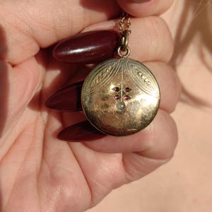 Peut inclure: Un collier avec un médaillon doré. Le pendentif rond présente un motif orné avec de petites pierres rouges et une perle centrale. La chaîne est délicate et également dorée.