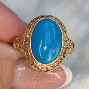May include: A gold ring featuring an oval, bright blue gemstone. The ring has an ornate, filigree design around the stone and on the band. The ring is a classic piece of jewelry.