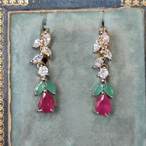 Può includere: Un paio di orecchini pendenti con una pietra preziosa di rubino, pietre di colore verde smeraldo e diamanti trasparenti. Gli orecchini sono dorati e hanno un design floreale delicato.