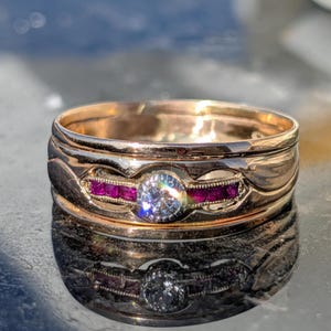 Op de afbeelding: Een gouden ring met een centrale, ronde, heldere diamant en twee rijen kleine, rode edelstenen. De ring heeft een uniek, gelaagd ontwerp met een reflecterend oppervlak, dat de ingewikkelde details en het vakmanschap laat zien.