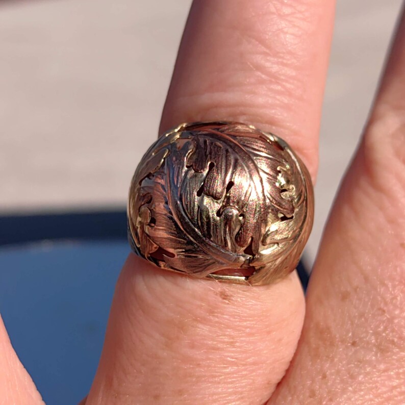 May include: A gold ring with a detailed leaf design. The ring is made of two-toned gold, with a darker gold band and a lighter gold leaf design.