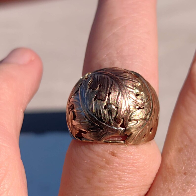 May include: A gold ring with a detailed leaf design. The ring is made of two-toned gold, with a darker gold band and a lighter gold leaf design.
