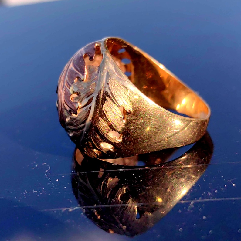 May include: A gold ring with a detailed, cut-out design of a feather. The ring is wide and has a rounded top.
