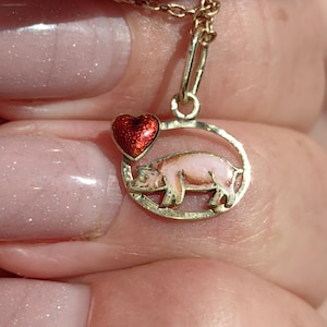 Puede incluir: Colgante dorado con un cerdo rosa dentro de un marco circular y un corazón rojo. El cerdo tiene un cuerpo rosa claro y detalles rosa más oscuros. El colgante está sujeto a una cadena dorada.